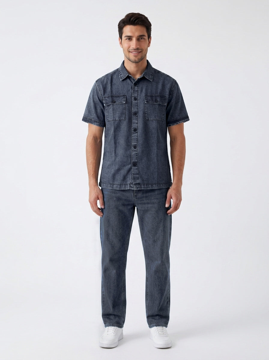 A man modeling a dark blue denim short-sleeve button-up shirt and matching dark blue denim jeans, paired with white sneakers, against a plain light gray studio background.