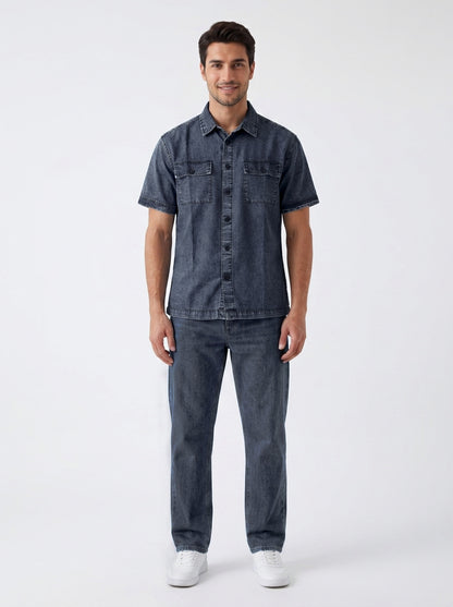 A man modeling a dark blue denim short-sleeve button-up shirt and matching dark blue denim jeans, paired with white sneakers, against a plain light gray studio background.