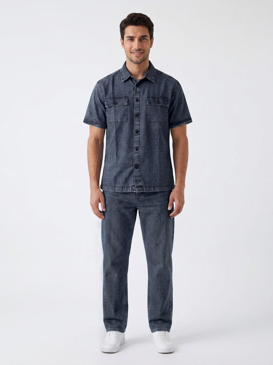 A man modeling a dark blue denim short-sleeve button-up shirt and matching dark blue denim jeans, paired with white sneakers, against a plain light gray studio background.
