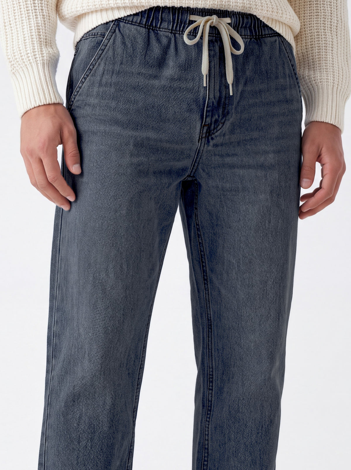 A close-up of a person wearing dark blue denim drawstring pants with a cream ribbed knit sweater, shown from the waist down against a plain white background.