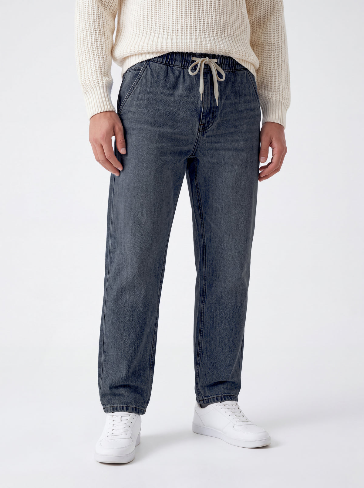 A person wearing dark blue denim jogger-style pants with an elastic drawstring waist, paired with a cream ribbed sweater and white sneakers, photographed against a clean light gray studio background.