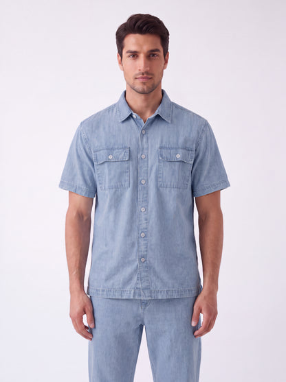 A man wearing a light blue short-sleeve button-up shirt and matching light blue jeans, standing against a plain white studio background.