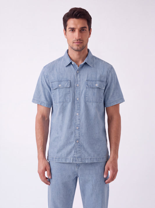 A man wearing a light blue short-sleeve button-up shirt and matching light blue jeans, standing against a plain white studio background.