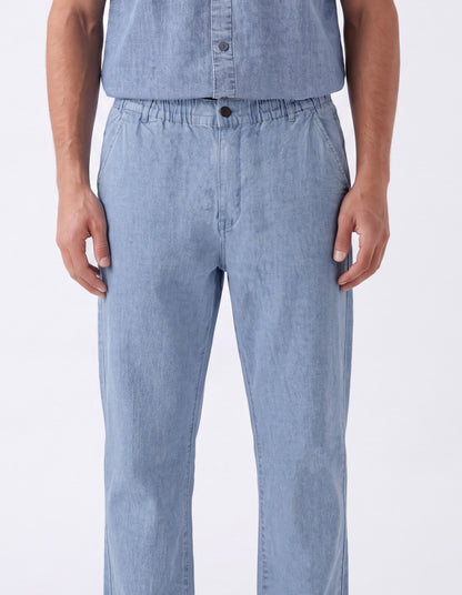 A person wearing light blue denim pants with an elastic waistband and a matching light blue button-up shirt, shown from the chest down against a plain white studio background.