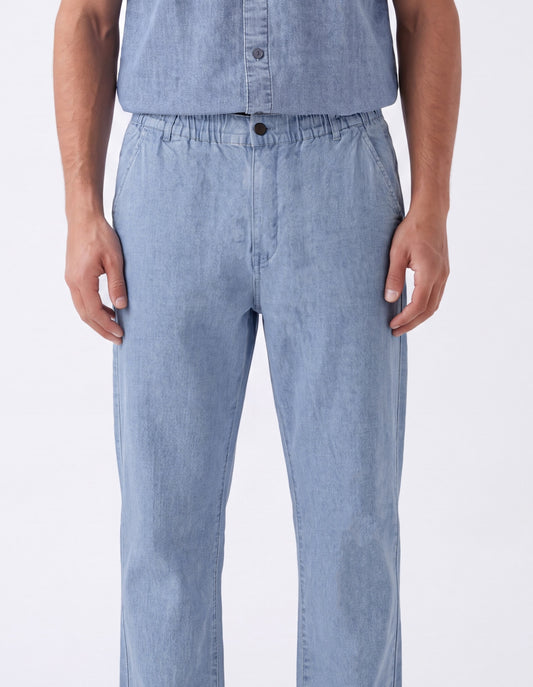 A person wearing light blue denim pants with an elastic waistband and a matching light blue button-up shirt, shown from the chest down against a plain white studio background.