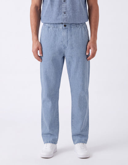 A person wearing light blue denim pants and a matching light blue button-front top, standing against a clean white studio background with white sneakers visible.