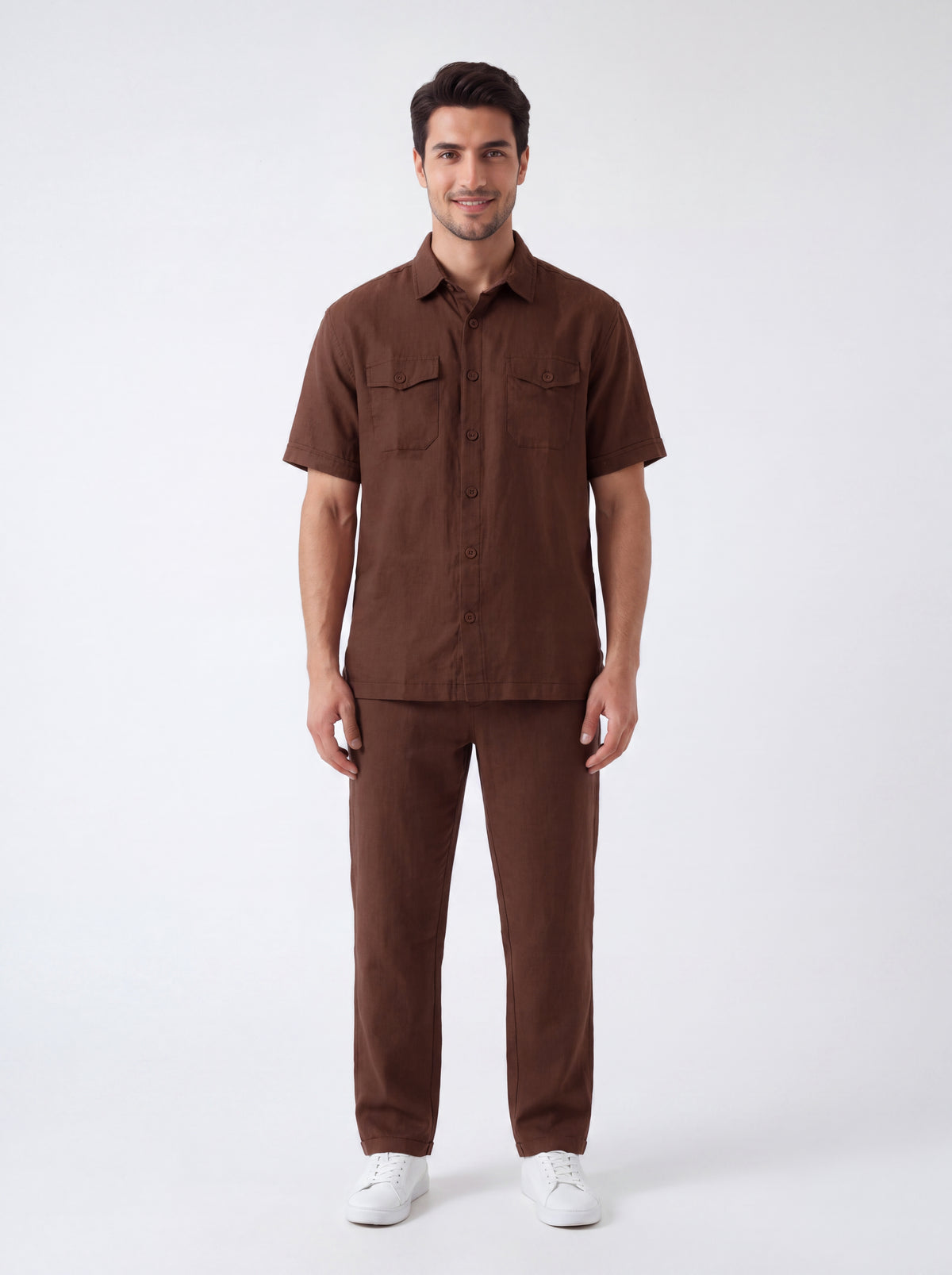 A man modeling a matching brown short-sleeve button-up shirt and trousers set, paired with white sneakers, against a plain light gray studio background.