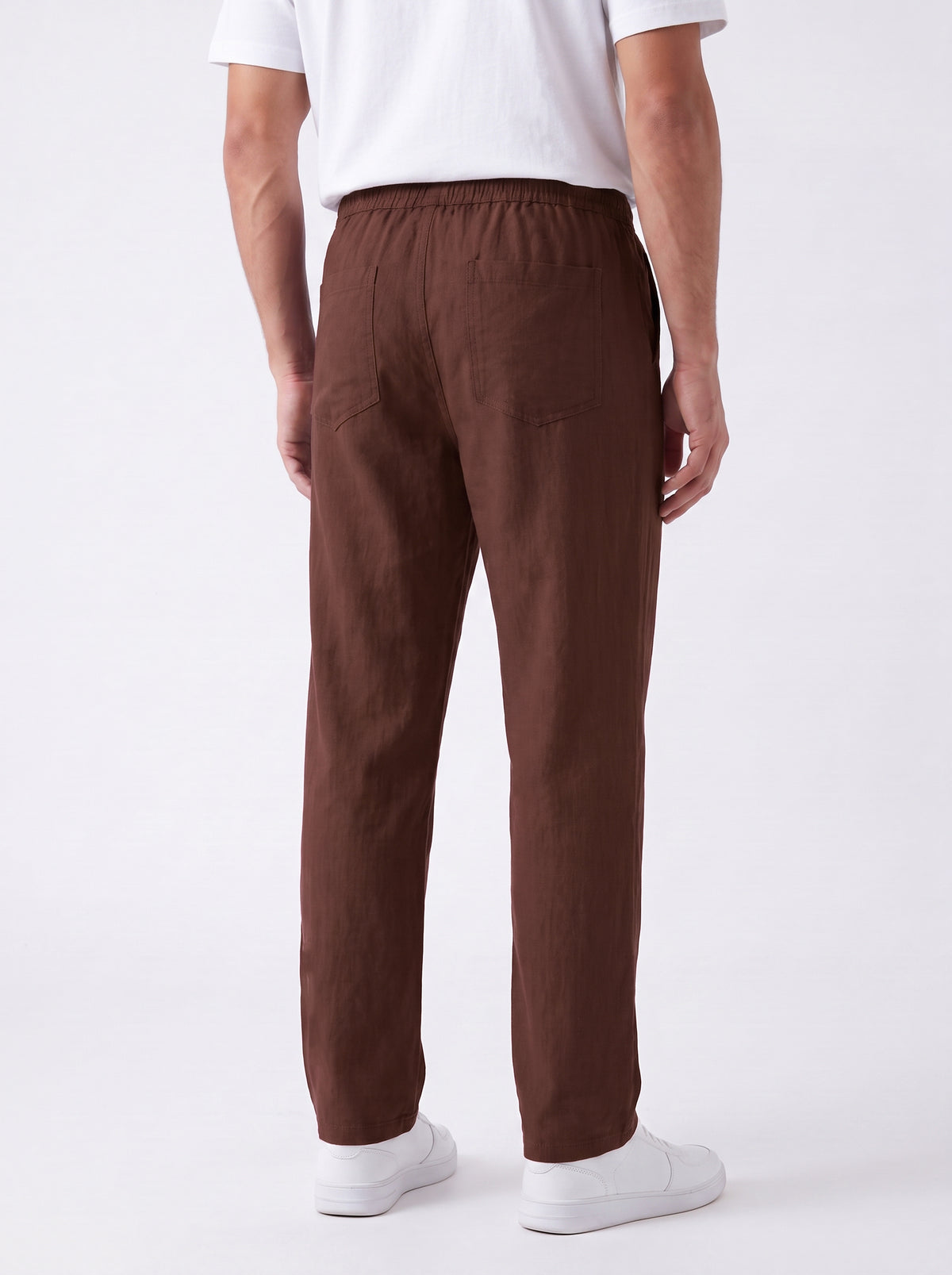 A man viewed from the back wearing brown straight-leg trousers, a white t-shirt, and white sneakers against a plain white studio background.