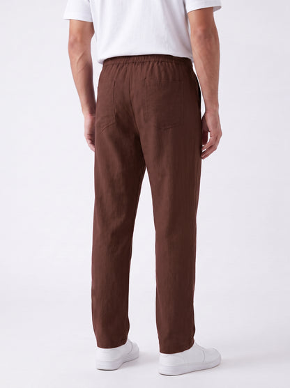 A man viewed from the back wearing brown straight-leg trousers, a white t-shirt, and white sneakers against a plain white studio background.