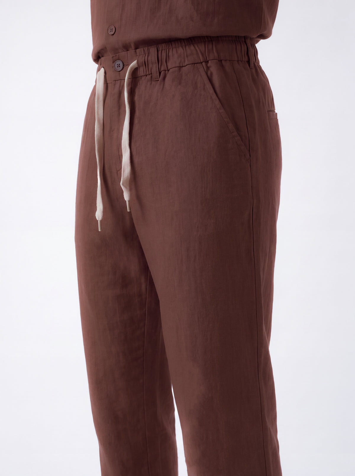 Close-up of a person wearing brown drawstring trousers with an elastic waistband and light beige drawstrings, shown against a plain light background.