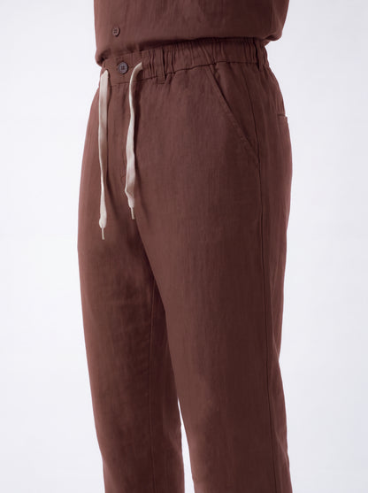 Close-up of a person wearing brown drawstring trousers with an elastic waistband and light beige drawstrings, shown against a plain light background.