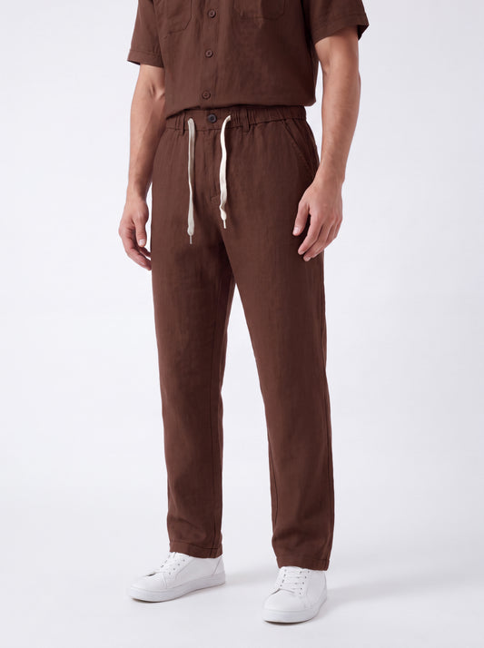 A person wearing brown drawstring pants and a matching brown short-sleeve top, paired with white sneakers, photographed against a clean light gray studio background.